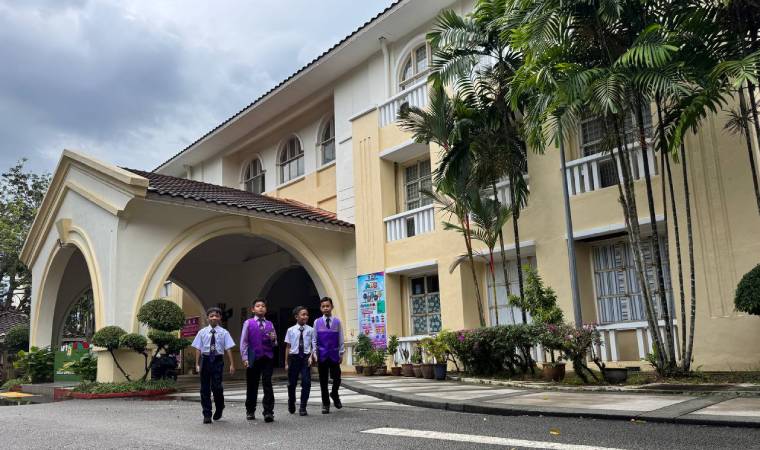 Cinco alunos uniformizados caminham em frente a prédio escolar de fachada clara com janelas arqueadas.