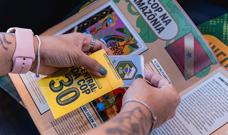 Mãos colam figurinhas com bandeiras no álbum Central da COP 30