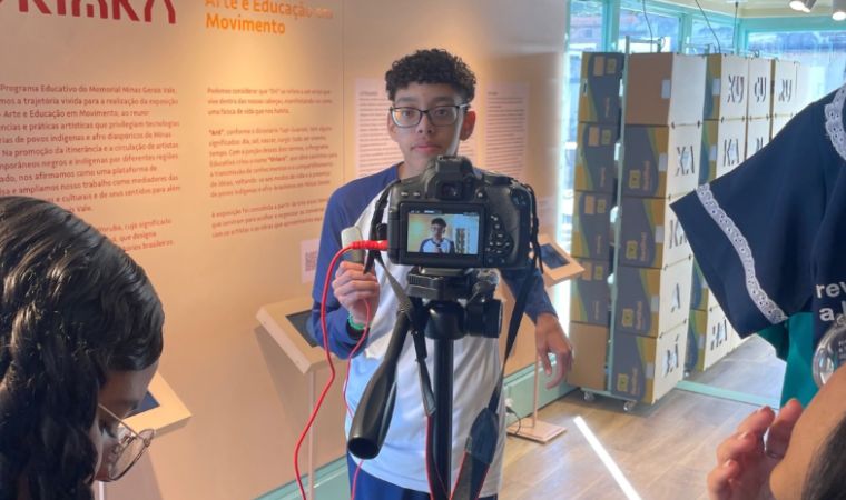 Jovem grava vídeo com câmera em frente a painel de exposição educativa