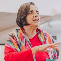 Mulher de cabelo curto fala gesticulando, vestida com roupa colorida