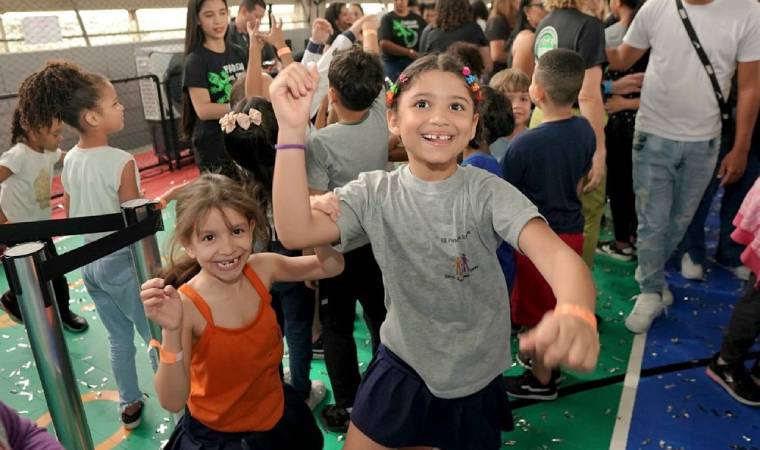 Duas meninas sorriem e dançam entre outras crianças em ambiente escolar
