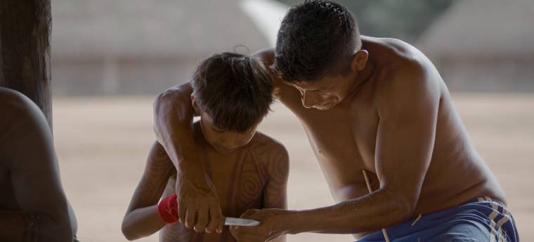 Homem indígena ensina menino a usar faca para talhar madeira