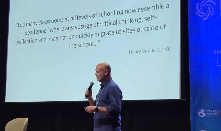 Michael Rowe fala ao microfone diante de telão com citação de Henry Giroux sobre educação