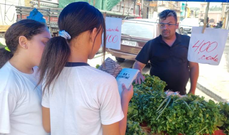 Duas meninas mostram um caderno a um vendedor de verduras em uma banca