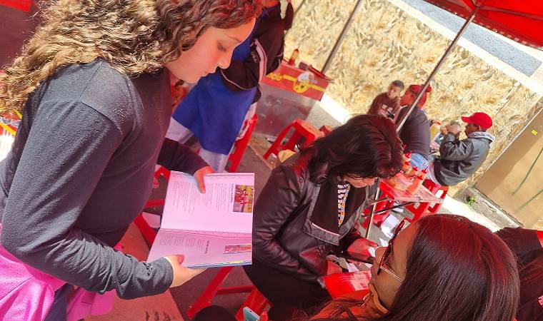 Menina segura um livro aberto enquanto conversa com outra criança em frente a barraca de pastel