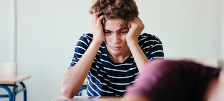 Adolescente com as mãos na cabeça, parecendo cansado ou frustrado, sentado em uma sala de aula
