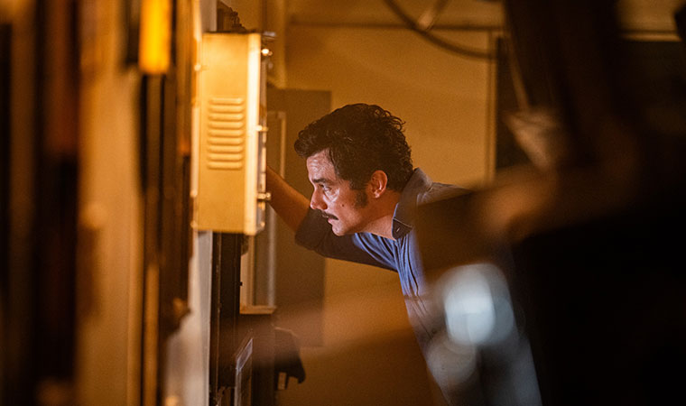 Wagner Moura inclinado observa equipamento em local escuro