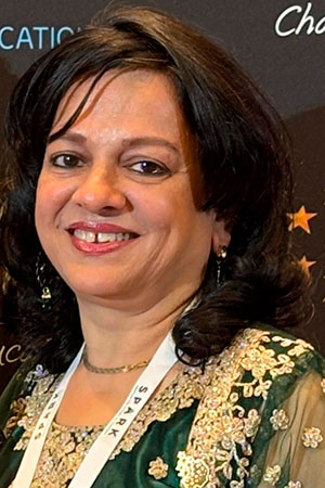 Vineeta Garg sorrindo e vestindo um vestido tradicional ou elegante, com bordados dourados e detalhes finos. Seu cabelo é escuro e levemente ondulado, e ela usa acessórios discretos. O fundo da foto parece ser uma parede escura com o logo de um evento ou organização, sugerindo que ela está em uma cerimônia ou celebração.