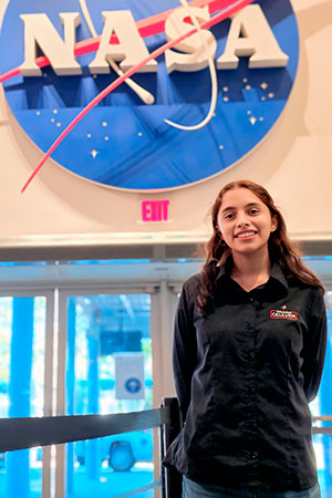 Valeria Palacios Cruz sorridente está posando em frente ao logotipo da NASA, com a marca visível em um grande círculo azul. Ela usa uma camisa preta com um logo visível (provavelmente de uma organização associada). O fundo mostra a entrada de um local, provavelmente uma instalação ou museu da NASA, o que sugere que ela está em uma visita ou trabalha ali.