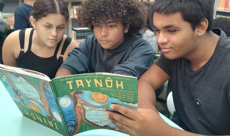 Três adolescentes leem juntos o livro infantil “Taynôh”
