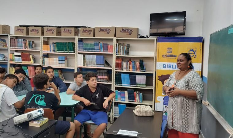 Professora em pé fala com estudantes sentados em biblioteca escolar