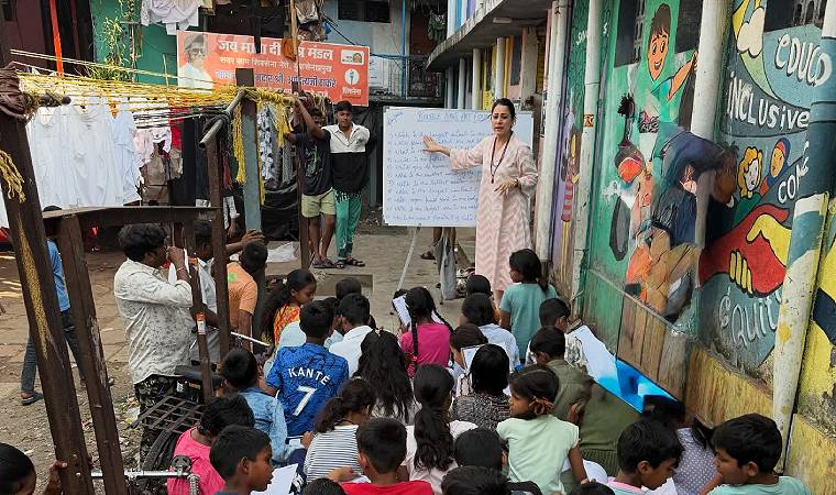 Rouble Nagi ensina grupo de crianças sentadas em viela com lousa ao fundo