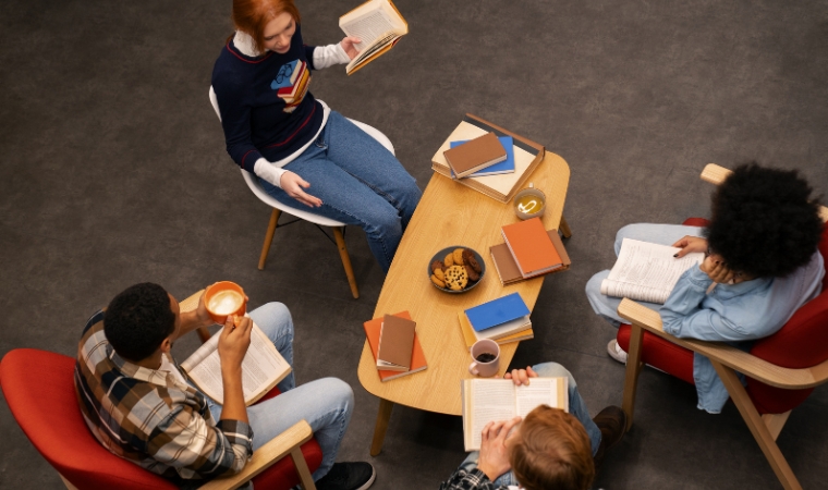 Estudantes sentados ao redor de mesa leem livros em atividade coletiva