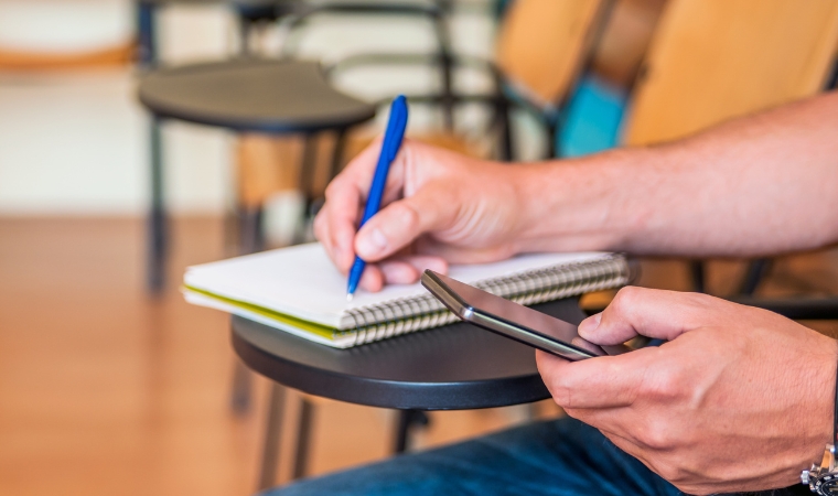 Pessoa usando smartphone e fazendo anotações em caderno em sala de aula, integrando tecnologia e estudo.